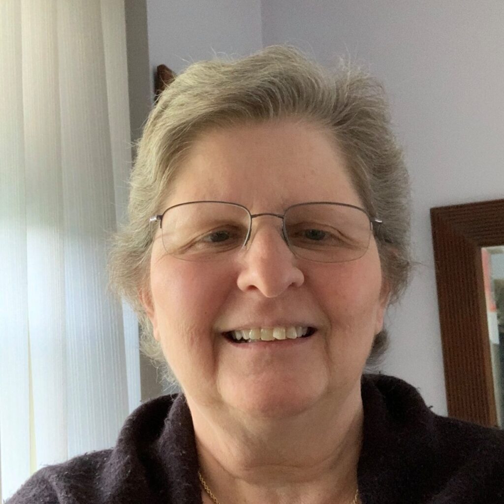 Photograph of Carol Schepper. A white woman wearing glasses with a thin silver metal frame inside a room with white walls and white vertical blinds. She is wearing a dark purple or brown sweater, and her hair is very short, gray, parted to the right. Her smile hints a bit of rambunctious mischief despite her overall practical no-nonsense vibe.