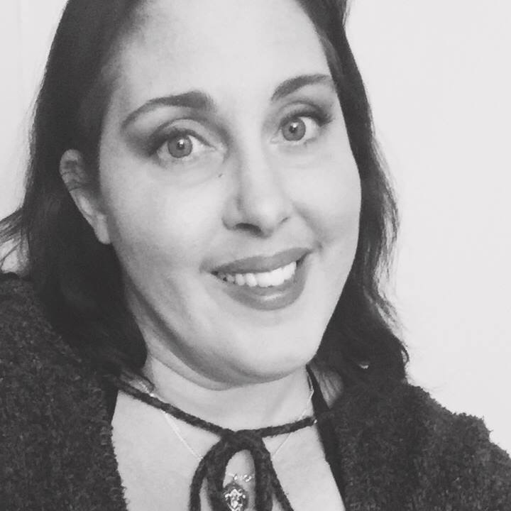 Photograph of Michelle Grewe. Black and white photo of a white woman in a gray sweater that ties across the top under the neck. She is wearing a silver pendant of Christ. Her dark brown hair sits just below her shoulders. Here wide eyes are looking to left but at you and her smile looks like the Cheshire Cat is trying not to smile.