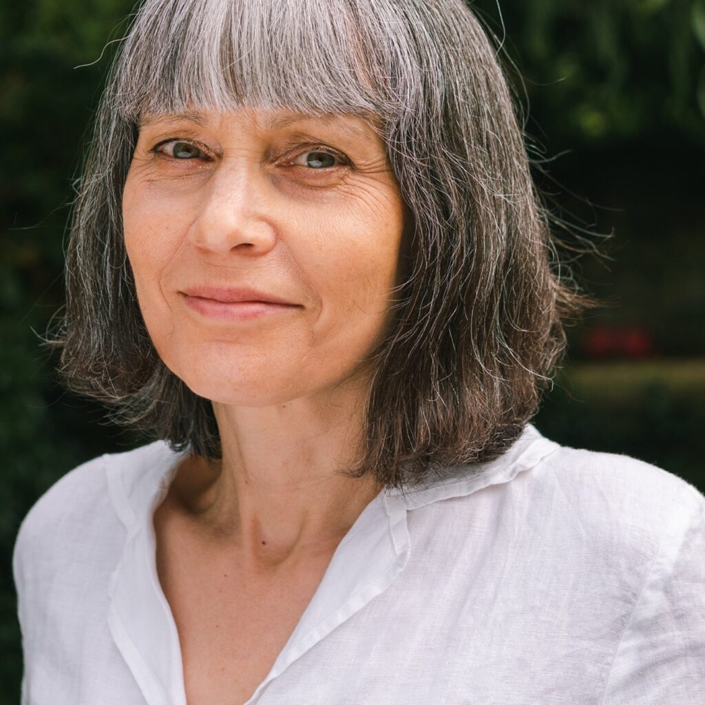 Photograph of Yvonne Spence. A woman with a brown bob and bangs with gray highlights. Her eyes are blueish with beautiful wisdom wrinkles like eye lines and face patterns on a bird. Her smile is soft and humbling. She's wearing a white button up blouse and standing in front of deep green trees and bushes in the background.